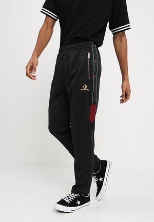 Persoon draagt zwarte Converse trainingsbroek met gouden logo, groene en rode zijstrepen, zwarte sneakers met witte veter en steraccenten.