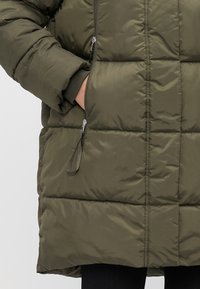 Mano metida en el bolsillo lateral de una chaqueta acolchada verde oliva con cremallera plateada visible y pantalones negros debajo.