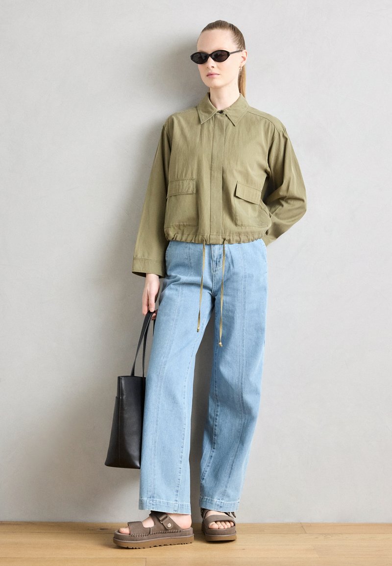 Oliwkowa krótka kurtka z dwoma przednimi kieszeniami, jasnoniebieskie szerokie jeansy, ciemnobrązowe sandały na platformie oraz czarna torba tote.