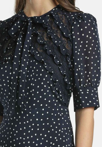 Marineblaues Kleid mit weißen Punkten, verziert mit Spitzenakzenten, Rüschen und einer Schleife am Ausschnitt; kurze, ballonförmige Ärmel.