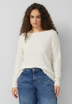 Femme aux cheveux bouclés portant un haut texturé blanc à manches longues et un jean bleu, debout devant un fond gris clair uni.