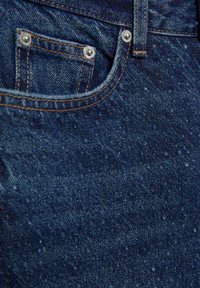 Tummansiniset farkut, joissa etutasku metallisten niittien kanssa, vaaleanruskeat saumat ja teksturoitu pinta, jossa on pieniä vesipisaroita.