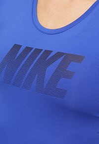 Modrý atletický top bez rukávů z hladké látky s velkým černým pruhovaným logem "NIKE" přes hrudník.