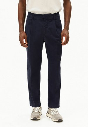 Homme portant un T-shirt blanc, un pantalon bleu marine foncé et des baskets gris clair, debout les bras détendus le long du corps devant un fond uni.