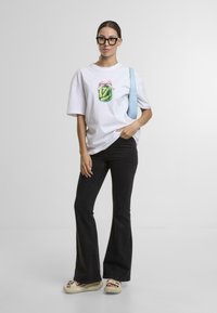 Bílý oversized tričko s barevným designem sklenice ve středu, v kombinaci s černými rozšířenými džíny a béžovými sandály. Modrá taška přes rameno.