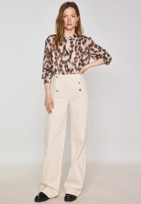 Blusa semitrasparente con stampa leopardata, con chiusura a bottone e maniche arrotolate, abbinata a pantaloni crema a vita alta e gamba larga con bottoni dorati.