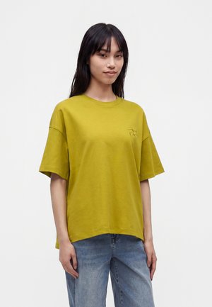 Jeune femme aux longs cheveux foncés porte une chemise ample jaune moutarde à manches courtes et un jean bleu sur un fond blanc uni.