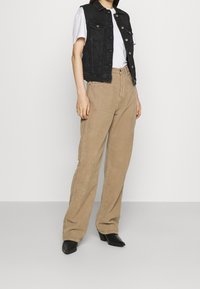Person som bär beiga kordflorsbyxor, svart ärmlös denimjacka, vit T-shirt och svarta klackade anklarstövlar, står mot en enkel vit bakgrund.