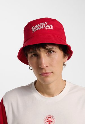 Ung person iført rød bøllehat med hvid tekst "DANISH DYNAMITE" og hvid skjorte med rødt emblem på ensfarvet baggrund.