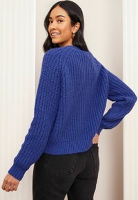 Mėlynai megztas džemperis su tekstūruota apdaila, su raštuotomis rankovėmis ir apatine dalimi. Dėvimas su aukštu juosmeniu juodomis kelnėmis, turinčiomis laisvą siluetą ir apvaliu iškirpimu.