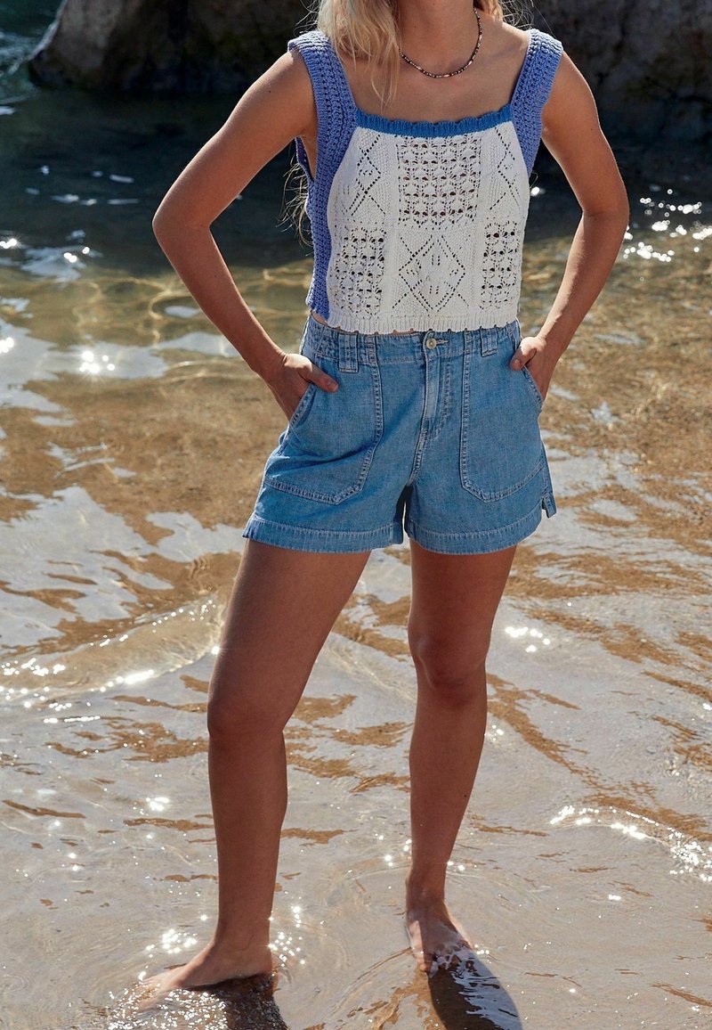Femme debout pieds nus dans l'eau peu profonde, portant un débardeur tricoté blanc et bleu et un short en jean taille haute, les mains dans les poches.