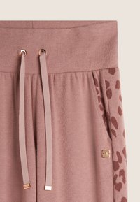 Pantalons de sport rose clair avec un motif léopard sur le côté, taille élastique avec cordon de serrage et accents en métal doré. Fini en tissu doux et texturé.