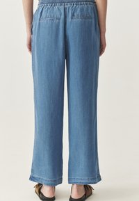 Heledad sinise denimi püksid, lõdva istuvuse, elastse vöökohaga ja tagakotiga. Tooriku serv pahkluu tasemel, koos lapikute sandaalidega.