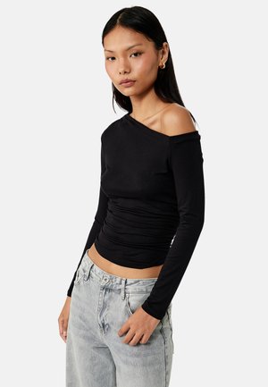 Vrouw draagt een lange mouw zwarte off-shoulder top en lichtgrijze high-waisted jeans, staand tegen een effen witte achtergrond.