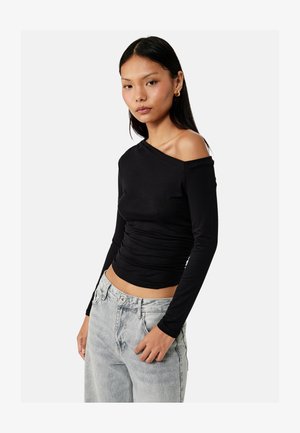 Vrouw draagt een lange mouw zwarte off-shoulder top en lichtgrijze high-waisted jeans, staand tegen een effen witte achtergrond.