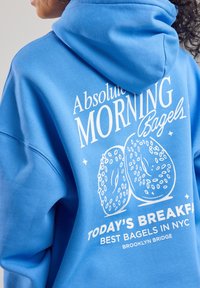 Blue hoodie featuring white text and bagel graphics. Material appears soft, with a front kangaroo pocket and a drawstring hood.