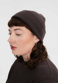 Donkerbruine gebreide beanie met een geribbelde textuur, iets schuin gedragen. De achtergrond is eenvoudig wit, waardoor de details van de muts worden benadrukt.