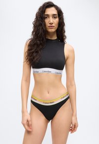 Γυναίκα με μακριά, σγουρά μαλλιά, που φοράει μαύρο crop top Calvin Klein με ψηλό λαιμό και μαύρα briefs που ταιριάζουν, με πολύχρωμη ζώνη με το λογότυπο της μάρκας.