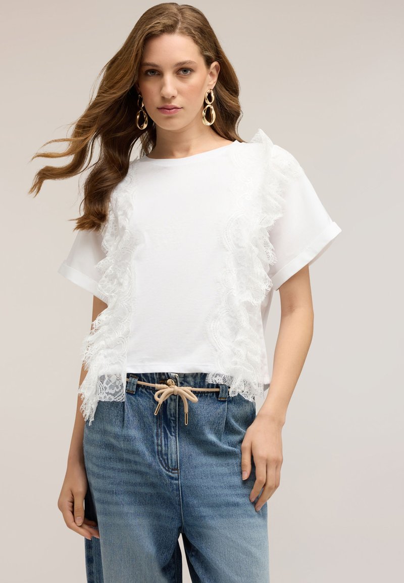 Woman with long brown hair wearing a white lace-trimmed short-sleeve top, gold hoop earrings, and high-waisted blue jeans with a beige belt.