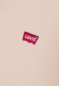 Riflesso del logo Levi's in rosso e bianco su uno sfondo in tessuto rosa chiaro, caratterizzato da una superficie testurizzata e un dettaglio di cucitura attorno all'etichetta.