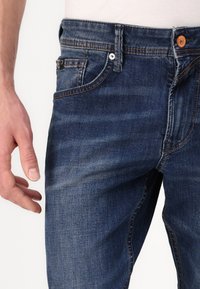 Jeans de mezclilla en azul oscuro con un ligero desgaste, diseño de cinco bolsillos, cierre estándar con cremallera y botón, con costuras en contraste de color naranja.