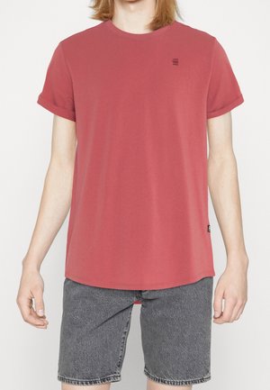 Personne portant un t-shirt rouge uni à manches courtes et un short en jean gris, debout devant un fond blanc.