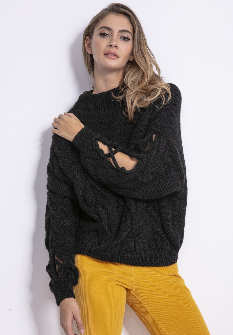 Czarny sweter z dzianiny z wzorem warkoczowym, z wycięciami na rękawach. Noszony z żółtymi spodniami z kordurowej tkaniny, które podkreślają kontrast faktur.