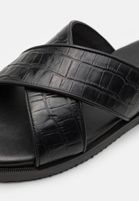 Sandales noires avec une sangle en cuir texturé à motif crocodile, présentant un design croisé et une semelle en caoutchouc lisse.