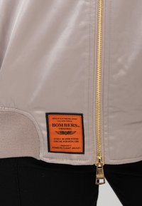 Beige bomberjack met gouden rits en oranje rechthoekige patch met het label "Bombers Original" nabij de zoom over een zwarte broek.