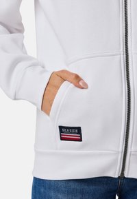 Weiße Zip-Hoodie mit einer Fronttasche, die ein schwarzes, rotes und weißes "SEASIDE" Logo-Patch auf der linken Seite hat. Weicher Stoff.