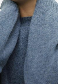 Närbild på en person som bär en mjuk, texturerad blå halsduk och matchande tröja, vilket visar halsen och den övre bröstområdet.