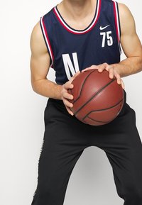 Laivastonsininen koripallopaita, jossa valkoinen "N"-kirjain ja numero "75", korostettuna punaisella ja valkoisella reunuksella, yhdistettynä mustiin urheiluhousuihin. Pelaaja pitelee koripalloa.