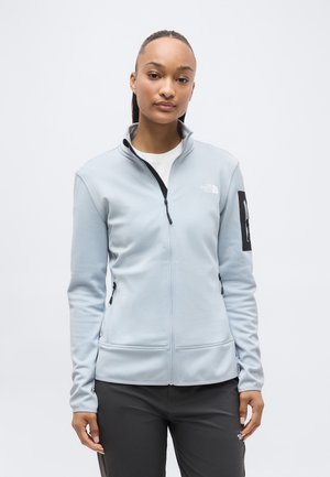 Młoda kobieta ubrana w jasnoszarą kurtkę na zamek i czarne spodnie, stojąca na tle białego, jednolitego tła.