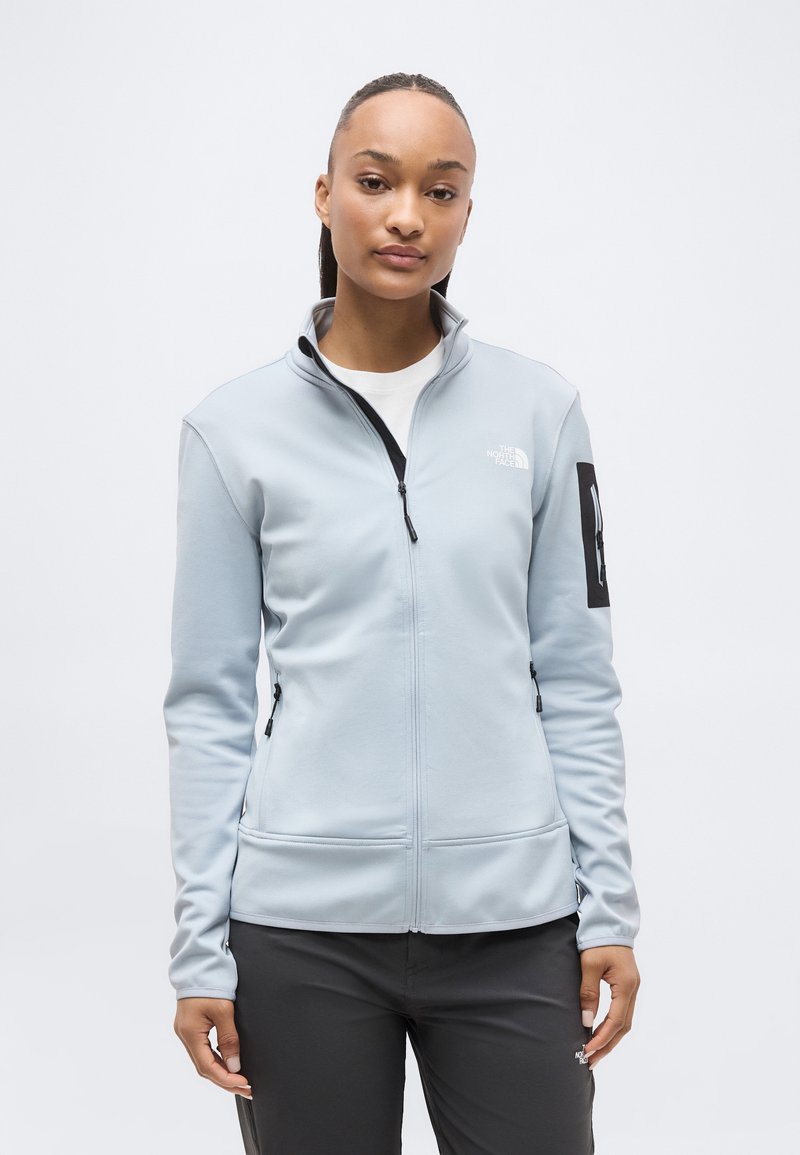 Νεαρή γυναίκα φορώντας ανοιχτό γκρι μπουφάν με φερμουάρ και μαύρο παντελόνι, στέκεται μπροστά σε απλό λευκό φόντο.