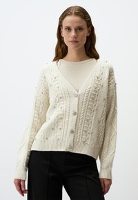 Crème cardigan met een v-hals, ingewikkeld kabelbreisel, versierd met kleine zilveren versieringen en geribde manchetten en zoom.