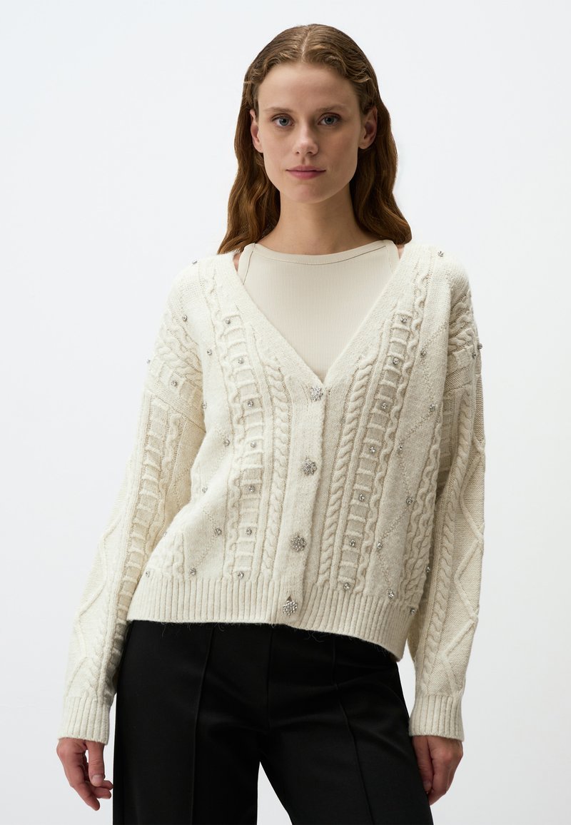 Crème cardigan met een v-hals, ingewikkeld kabelbreisel, versierd met kleine zilveren versieringen en geribde manchetten en zoom.