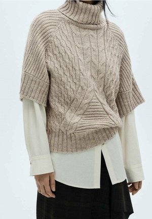 Beżowy sweter z krótkim rękawem i golfem o splocie warkoczowym, noszony na białej, długorękawowej koszuli, zestawiony z czarnymi spodniami.