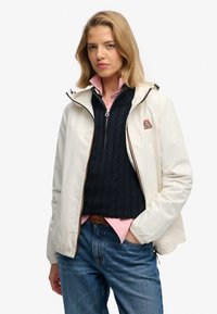 Biała kurtka z kapturem i zamkami błyskawicznymi, z pomarańczowymi akcentami, nałożona na granatowy sweter w warkocze i różową koszulę z kołnierzykiem, noszona z niebieskimi dżinsami.