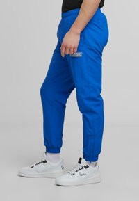 Blue athletic pants with elastic waistband, elastic cuffs, and logo detail; paired with white sneakers featuring black accents and textured soles.