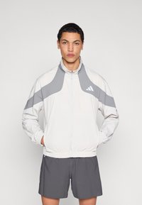 Lightweight white jacket with gray accents, featuring a high collar and elastic cuffs. Model pairs with gray shorts, highlighting sporty design.