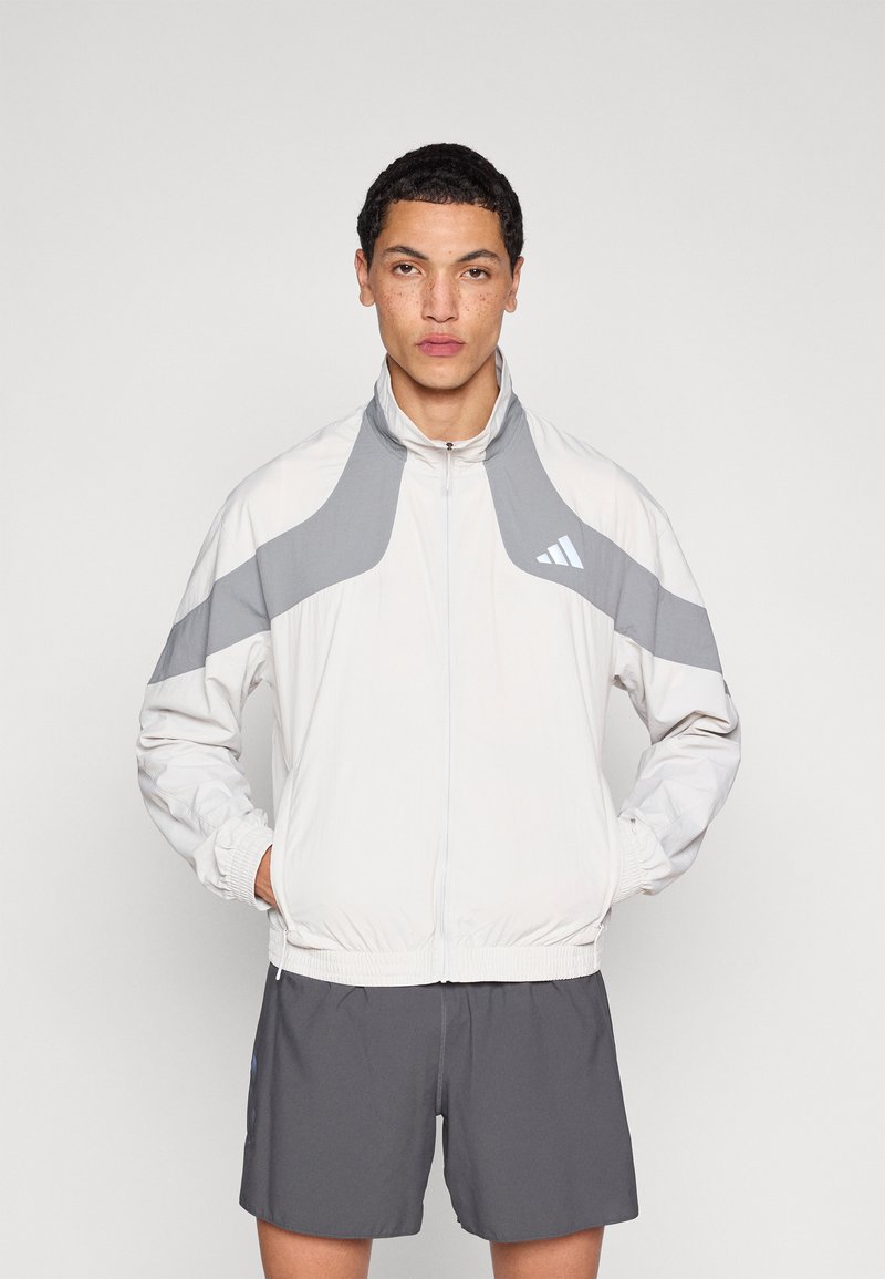 Lightweight white jacket with gray accents, featuring a high collar and elastic cuffs. Model pairs with gray shorts, highlighting sporty design.