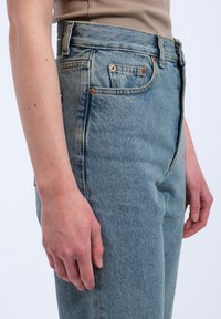 Light blue high-waisted jeans made of denim. Features copper rivets and a front zipper. The fabric has a slight texture and faded appearance.
