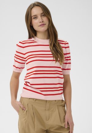 Tânără cu păr lung și castaniu, purtând un top cu mâneci scurte în dungi roz și roșu și pantaloni bej, stând cu o mână în buzunar.