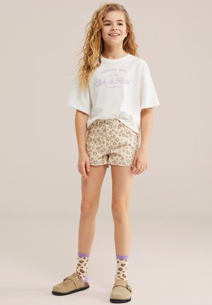 Fille aux longs cheveux bouclés portant un t-shirt blanc à motif, un short imprimé léopard, des chaussettes assorties et des chaussures beiges sans lacets, debout et souriante.