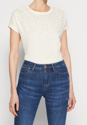 Kobieta ubrana w biały top z krótkim rękawem ozdobiony małymi ćwiekami na górnej części piersi, w połączeniu z niebieskimi, wysokim stanem jeansami.