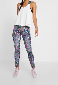 Vrouw draagt een wit mouwloos topje, kleurrijke legging met bloem- en abstracte patronen, en lichtroze sportschoenen.