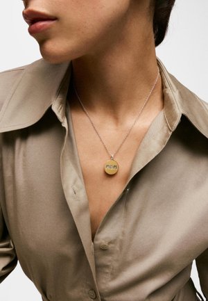 Zbliżenie osoby noszącej beżową koszulę na guziki i srebrny naszyjnik z okrągłą złotą zawieszką z grawerem "MOM".