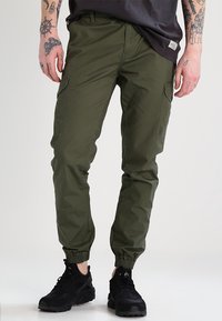 Olivgröna cargobyxor i en bomullsblandning, med elastiska muddar, flera sidofickor och en avslappnad passform. Matchas med svarta sneakers.