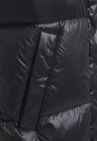 Detail eines schwarzen Daunenjacke, die eine glatte, glänzende Textur mit genähten Abschnitten und einer seitlichen Reißverschlusstasche aufweist.