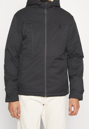 Veste noire à capuche avec fermeture éclair, deux poches poitrine verticales zippées, portée avec un pantalon clair, mains détendues le long du corps.